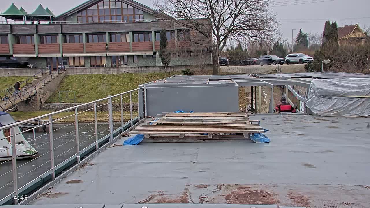 A gray flat roof with wooden pallets sits atop a houseboat next to a canal, overlooking a building and overcast sky.