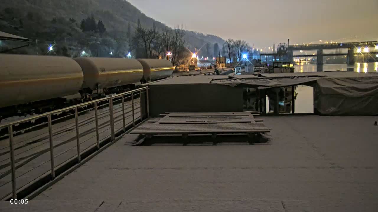 A snow-covered barge is situated near a train carrying tanker cars, all under a twilight sky next to a river and bridge.