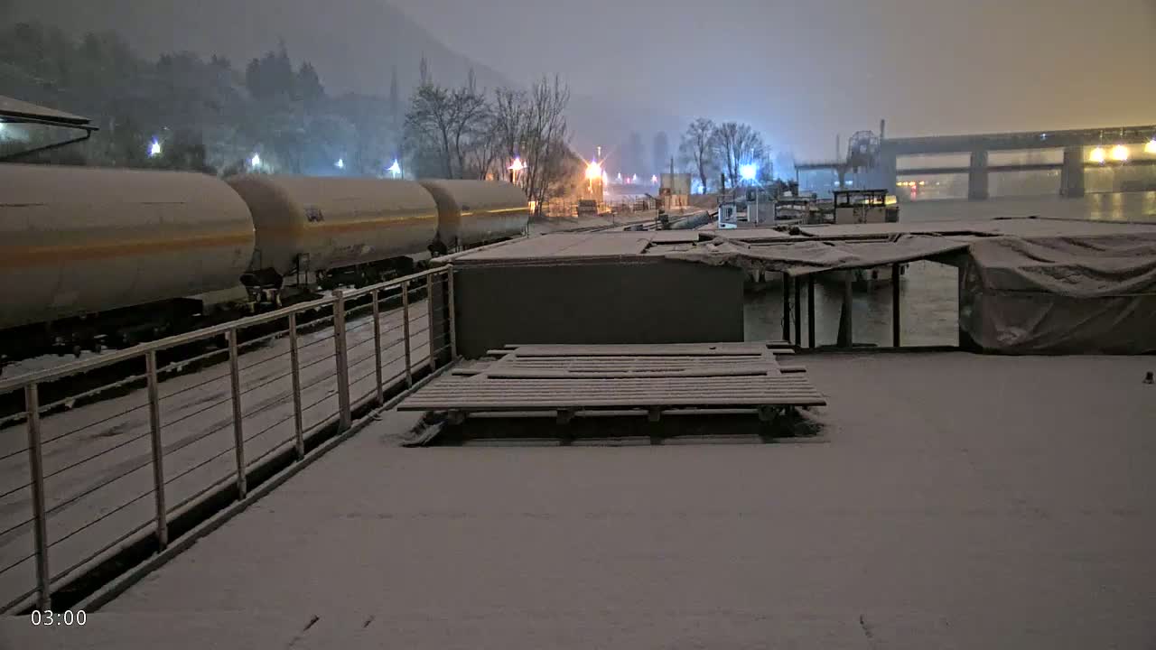 Snow covers a riverbank at night, where a train of tanker cars is parked next to a snow-covered barge and several other boats.