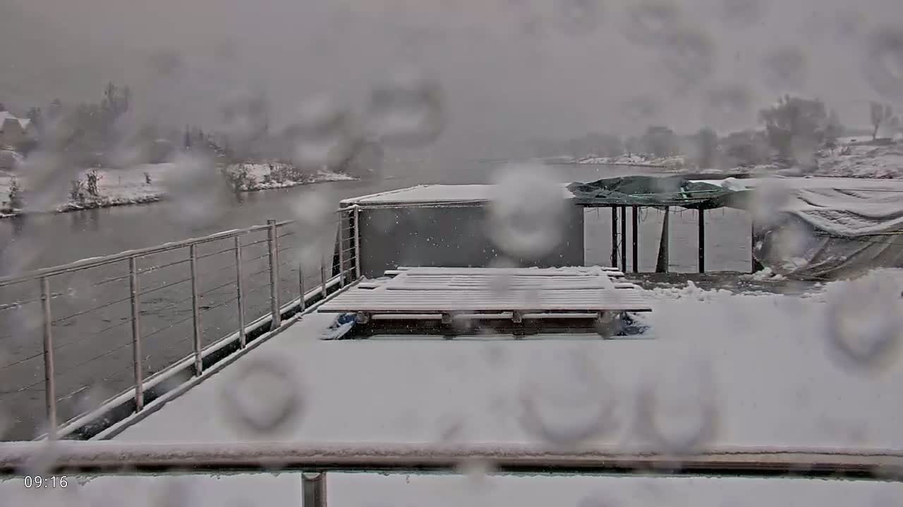 A snow-covered barge on a river is viewed through a rain-covered window during a snowfall.