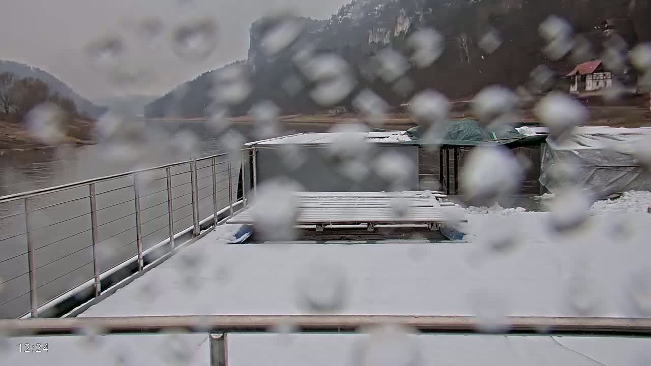 A snow-covered deck of a boat on a river, viewed through rain-covered glass, with hills visible in the background.