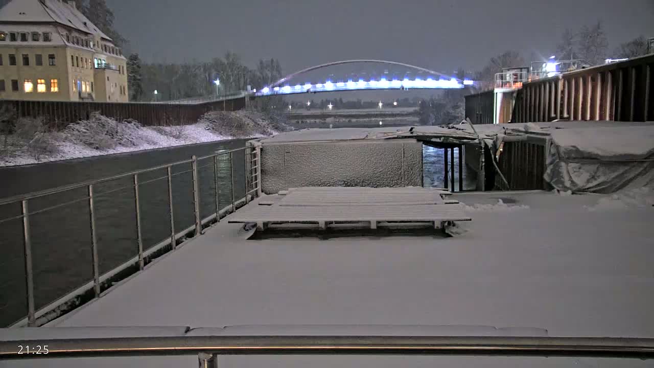 A snow-covered deck overlooks a snow-covered riverbank, a lit bridge, and a building at night.