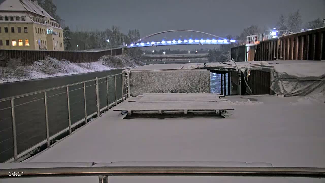 A snow-covered deck overlooks a snow-dusted canal, a lit bridge, and a building in the distance under a nighttime sky.
