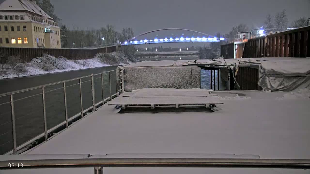 A snow-covered deck overlooks a snow-covered riverbank, a lit bridge, and a building in the distance under a nighttime sky.