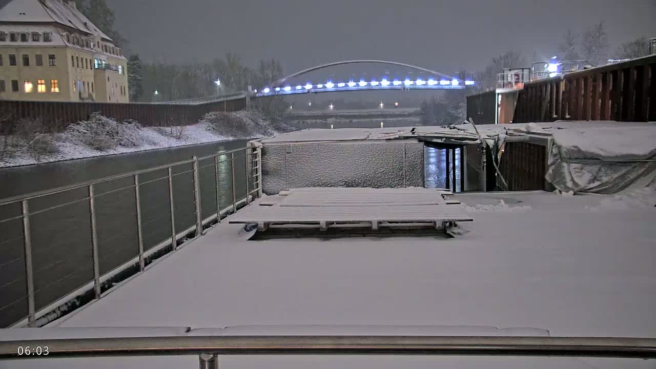 A snow-covered barge is moored on a canal at night, with a lit bridge in the background.