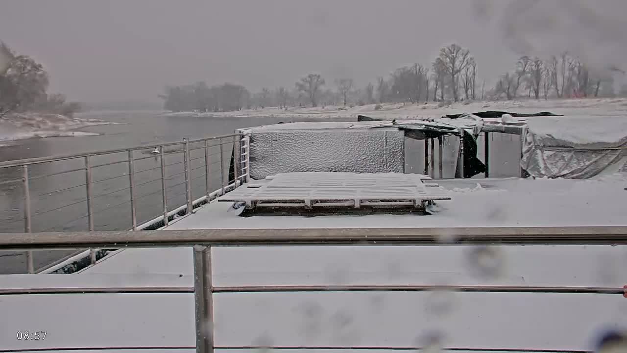 A snow-covered barge deck with metal railings overlooks a river during a snowfall.