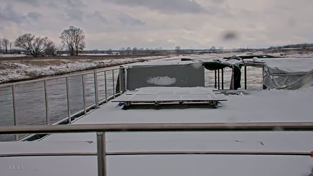 A snow-covered barge is docked on a river, with a snowy landscape visible in the background under a cloudy sky.