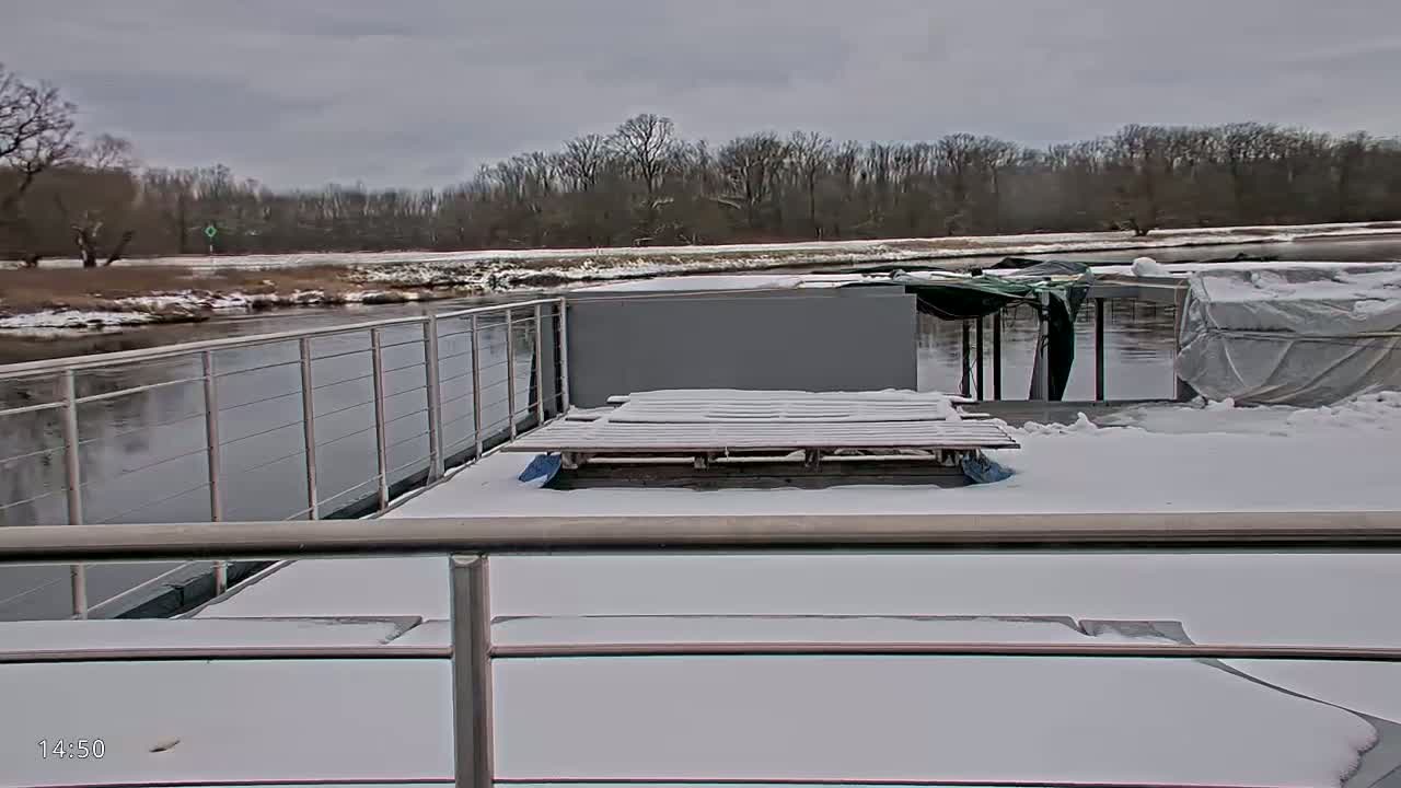 A snow-covered deck of a boat overlooks a calm river bordered by a snowy, wooded shoreline under an overcast sky.