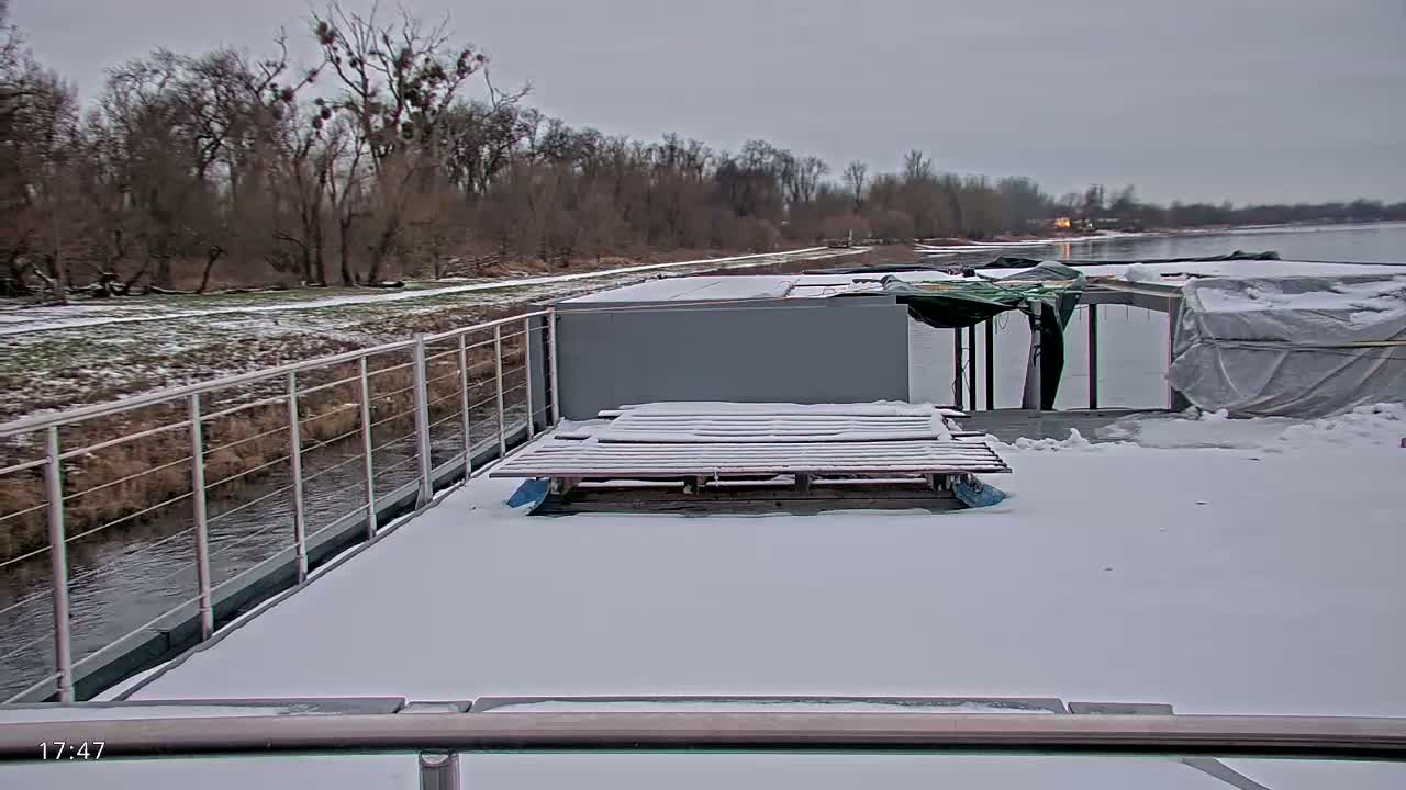 A snow-covered barge deck with a wooden pallet and a view of a snow-dusted riverbank and trees under an overcast sky.