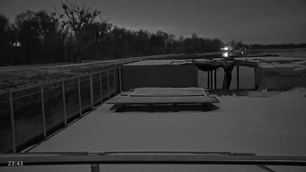 A snow-covered deck with a pallet and a metal fence overlooks a dark, still body of water under a night sky.