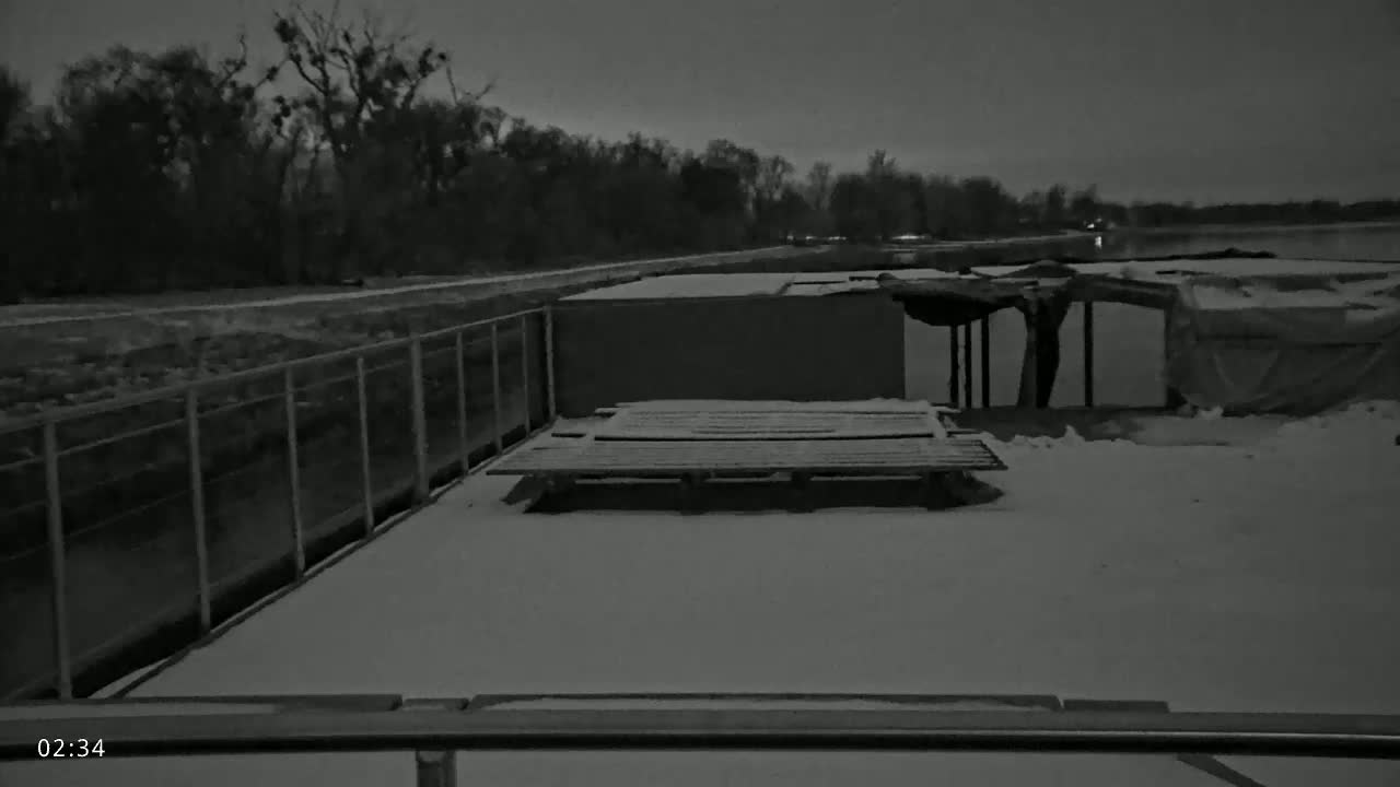 A snow-covered platform with a wooden pallet sits near a dilapidated structure and a metal fence bordering a dark body of water under a twilight sky.
