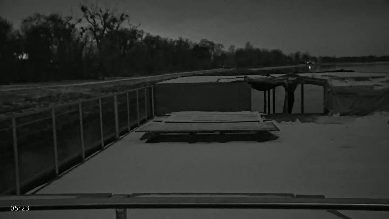 A snow-covered platform with a metal grate is seen next to a dilapidated structure and chain-link fence under a dark night sky.