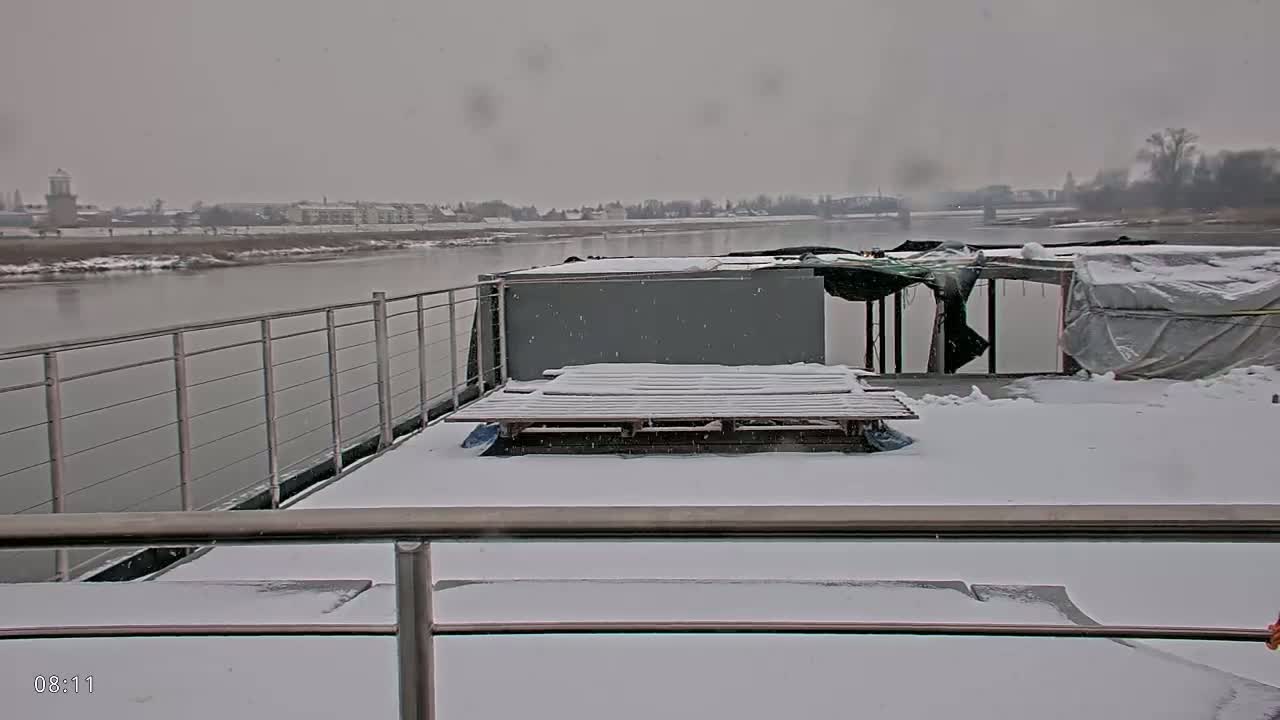 A snow-covered barge deck overlooks a wide river and a distant town under a light snowfall.