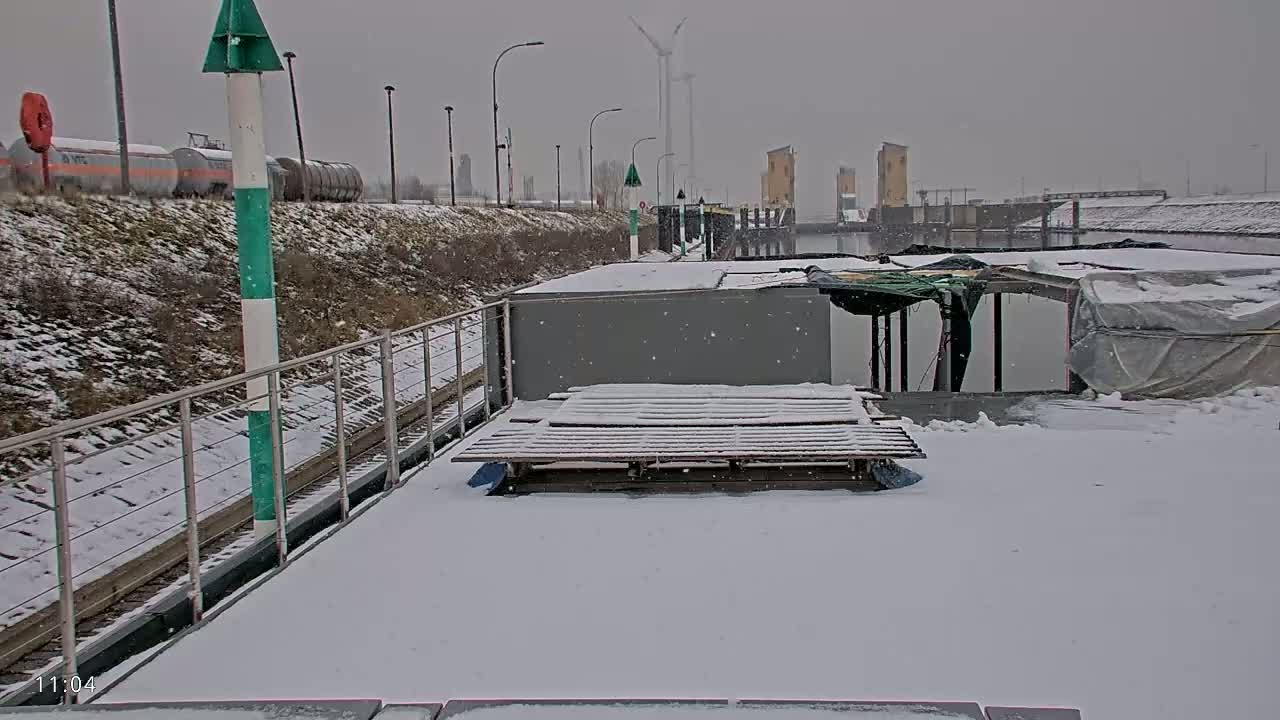 A snow-covered barge is docked at a canal, with light snow falling, and railway tracks and industrial buildings visible in the background.