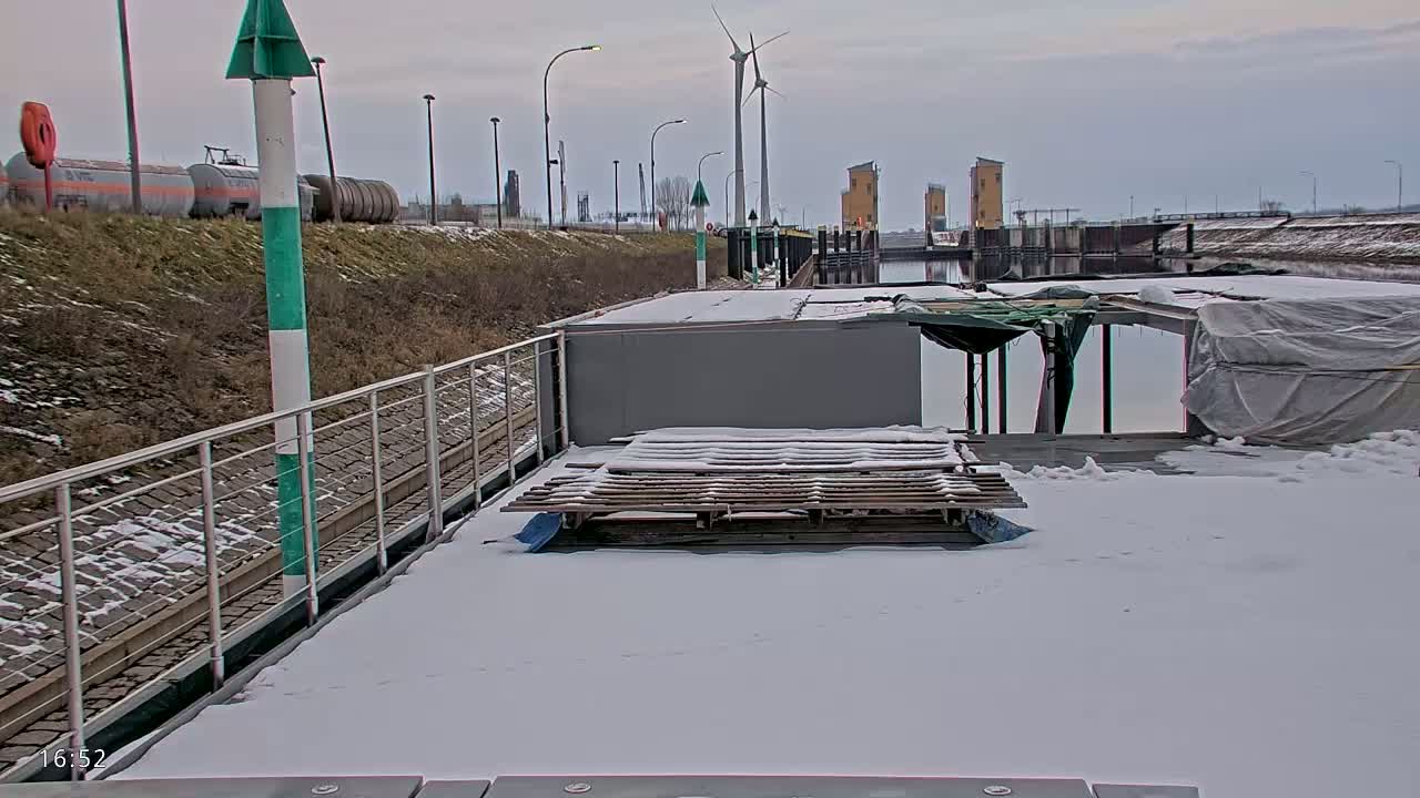 A snow-covered barge is docked at a canal, with a wind turbine visible in the distance under an overcast sky.