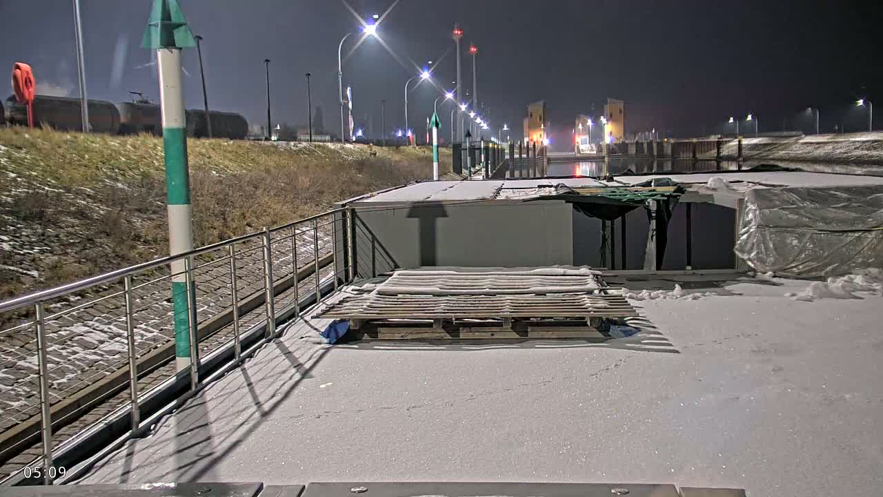 A snow-covered barge deck at night, with a pallet of wood on the deck, overlooks a canal lit by streetlights.