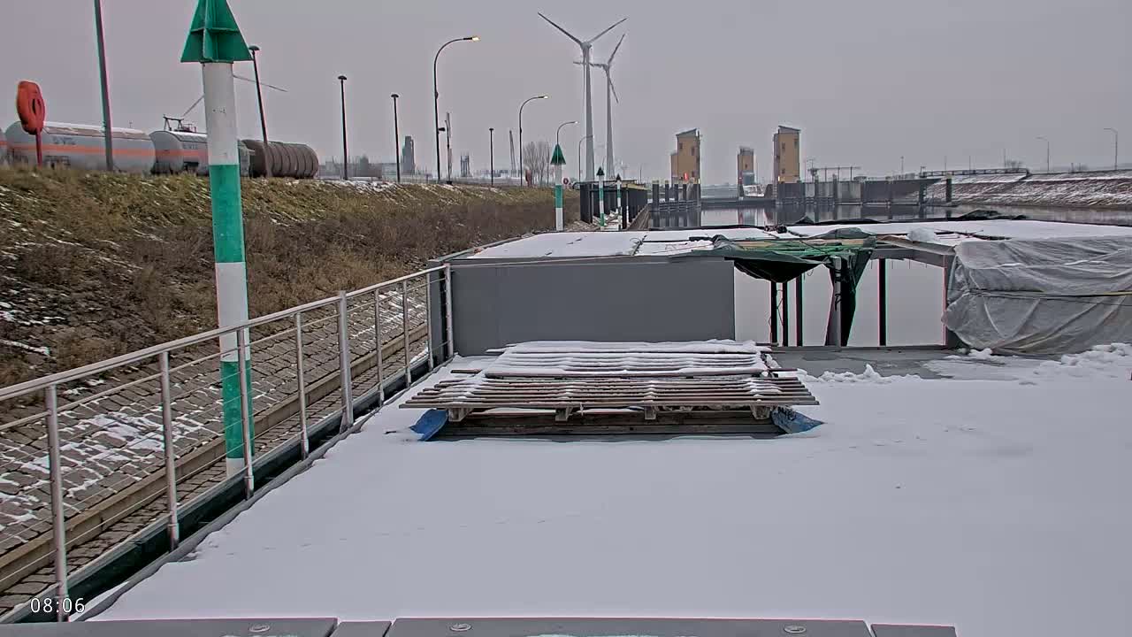 A snow-covered barge is docked at a canal, with a wind turbine and industrial buildings visible in the background under an overcast sky.