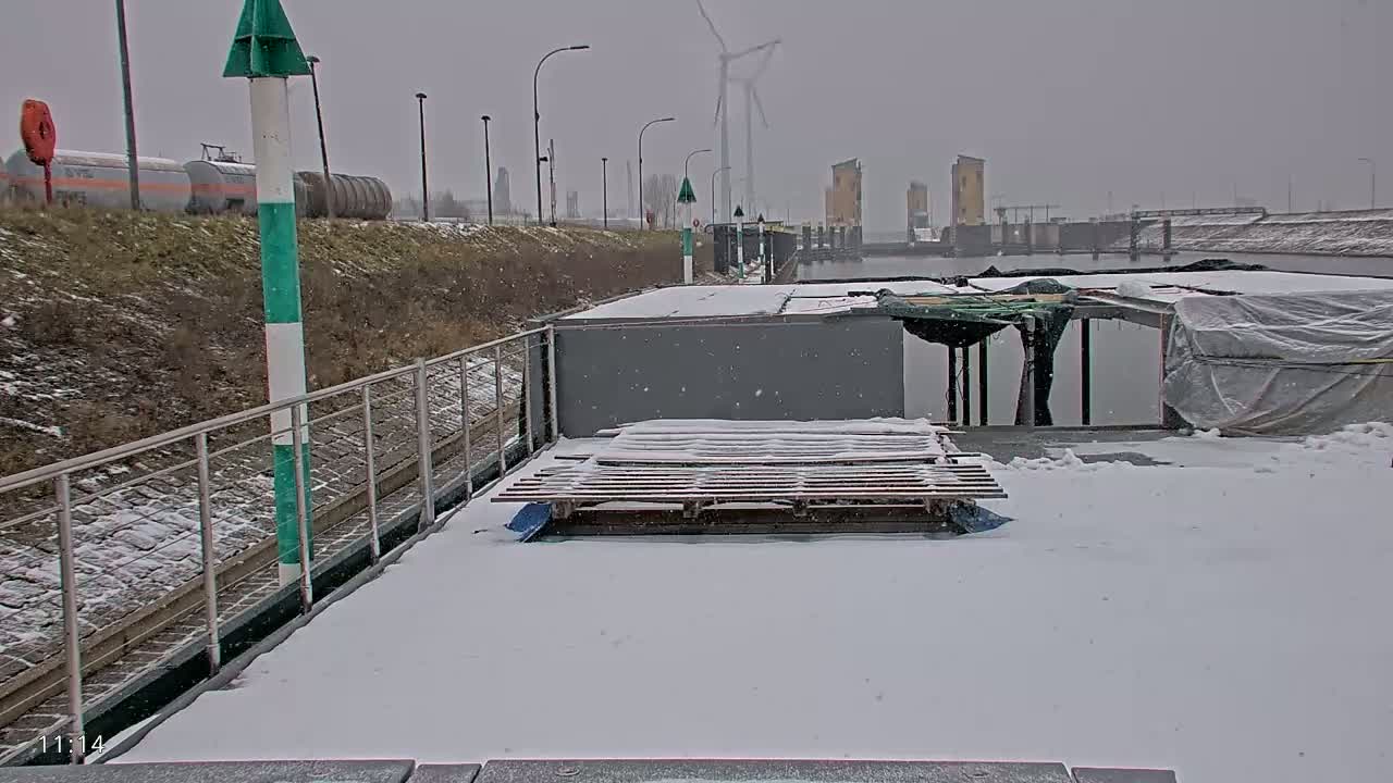 A snow-covered barge is moored at a canal, with a wind turbine and industrial buildings visible in the distance under a light snowfall.