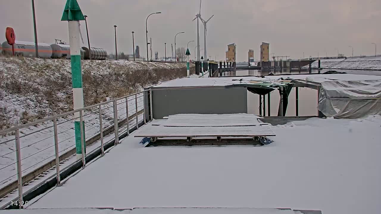 A snow-covered barge sits near a waterway with a wind turbine and lock system visible in the background under an overcast sky.