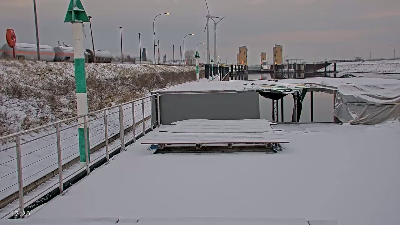 A snow-covered dock with a gray structure and wooden planks sits beside a waterway near a lock system under an overcast sky.