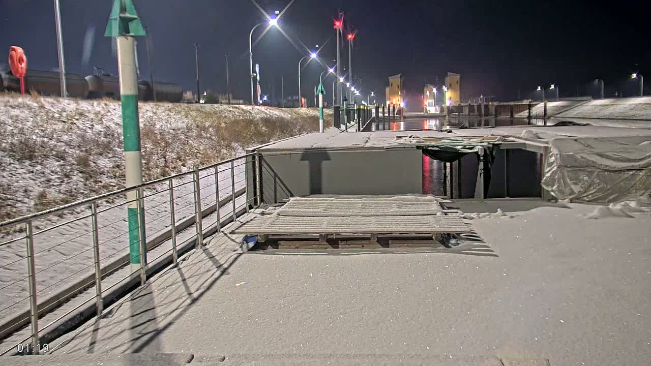 A snow-covered barge sits at a nighttime dock beside a canal, with lights illuminating the waterway and surrounding area.