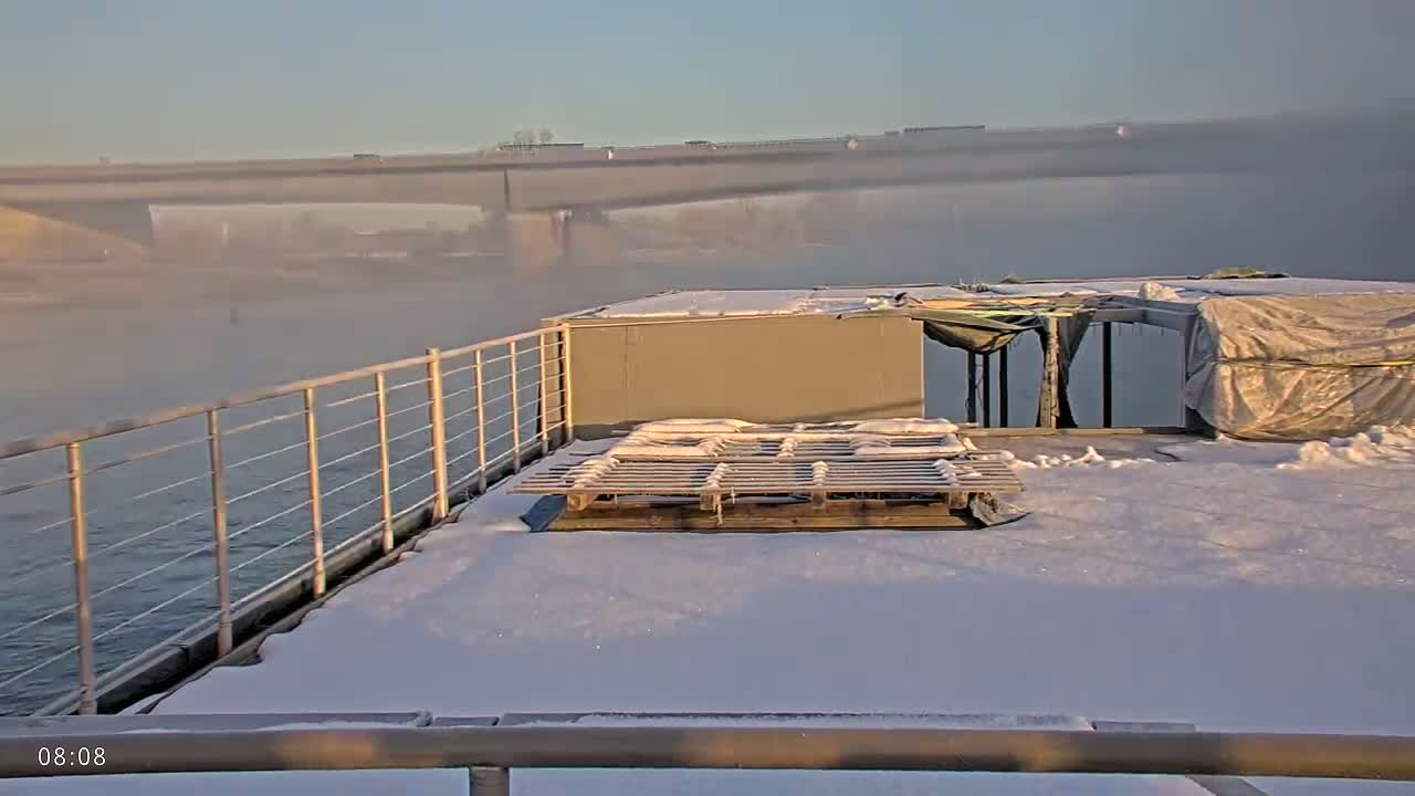 A snow-covered deck of a boat on a foggy river with a bridge in the background.