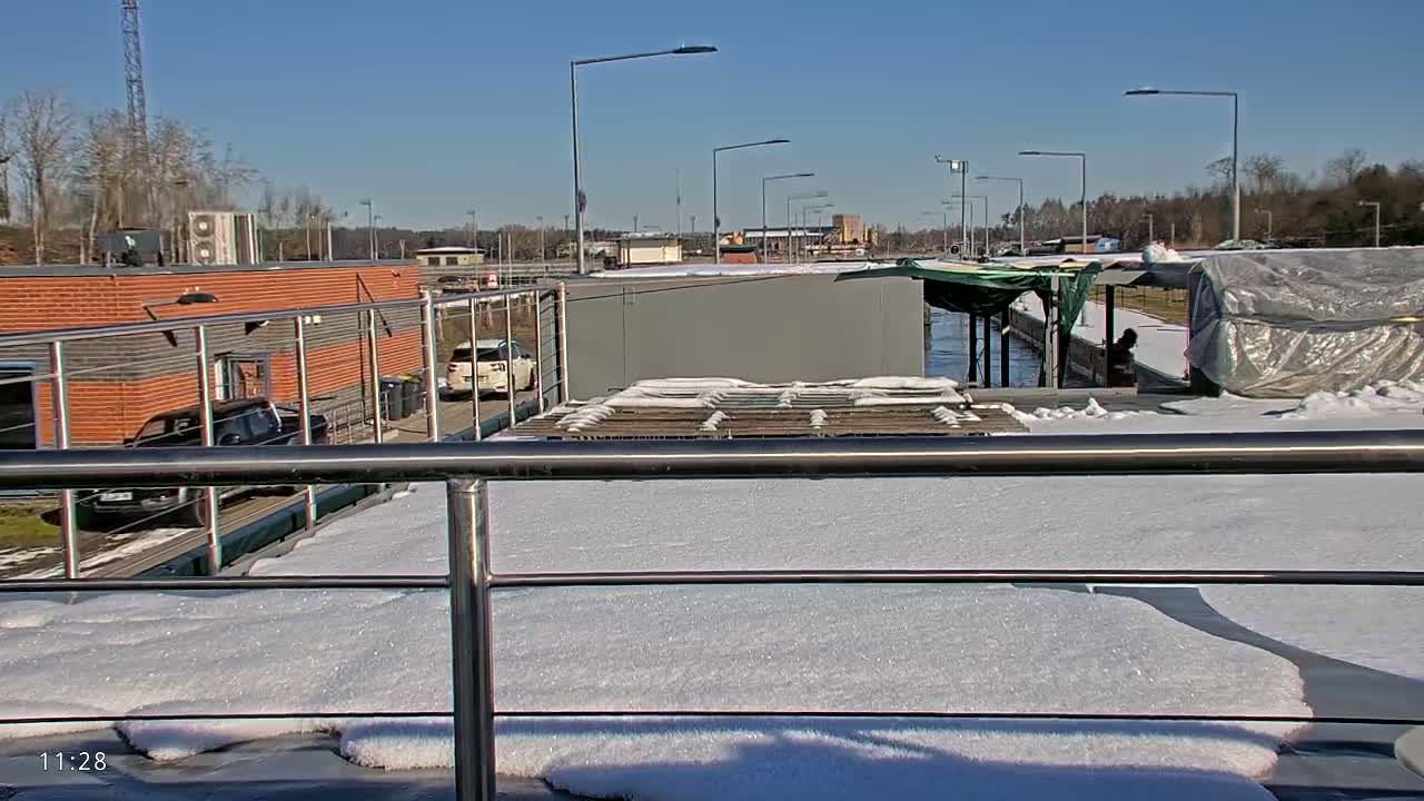 A snow-covered deck overlooks a canal, with buildings and vehicles visible in the background under a sunny sky.