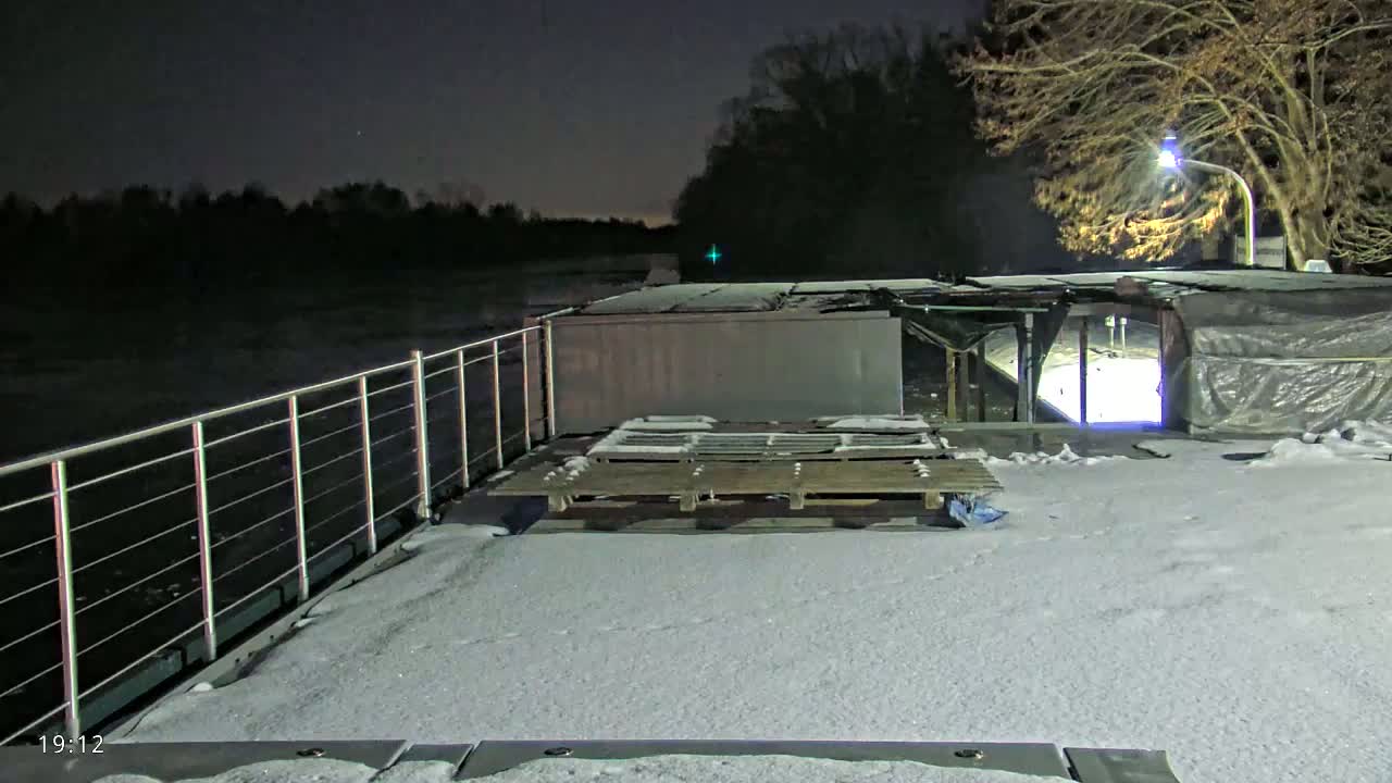 A snow-covered deck of a barge or boat at night, overlooking a dark river under a clear sky, with a bare tree and light visible in the background.