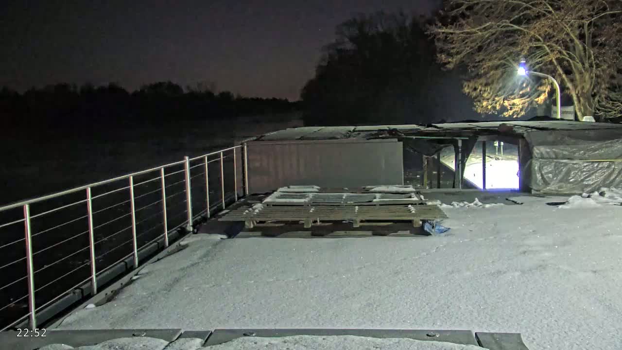 A snow-covered deck on a boat at night overlooks a dark river under a partly cloudy sky.