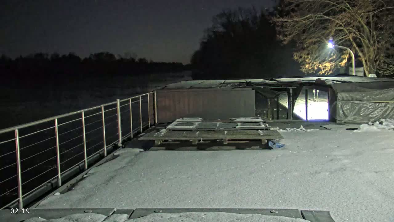 A snow-covered deck at night overlooks a dark body of water, with a metal railing and a pallet visible on the deck.
