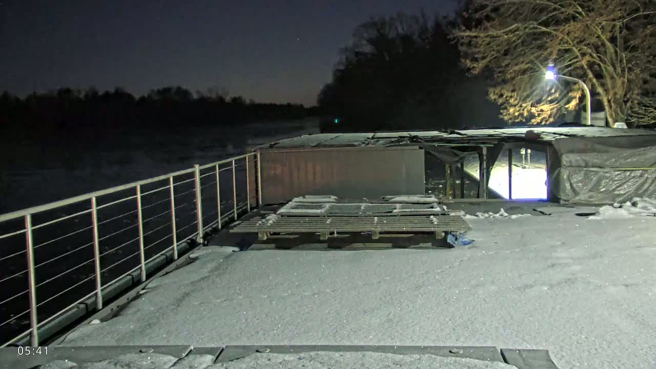 A snow-covered barge is moored at night next to a dark, still body of water, under a mostly clear sky, with bare trees visible in the background.