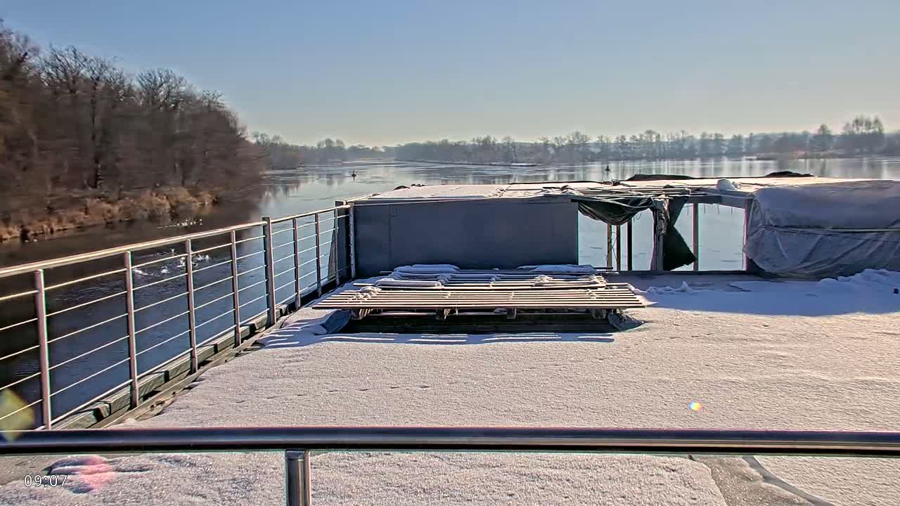 A snow-covered deck of a boat overlooks a calm river bordered by leafless trees under a clear, sunny sky.