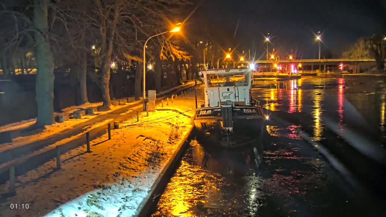 A small tugboat is moored at a snow-covered canal bank at night under streetlights.