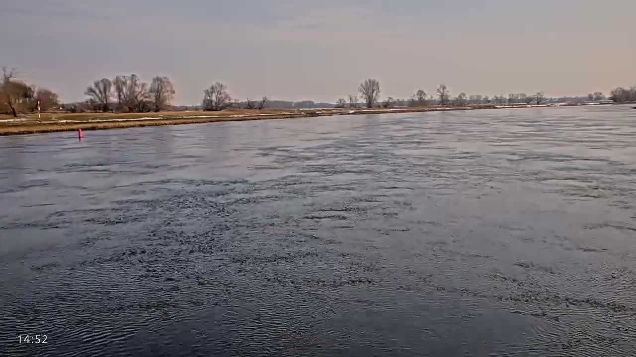 A wide, slightly choppy river flows under a pale, overcast sky past a flat, treeline bank.