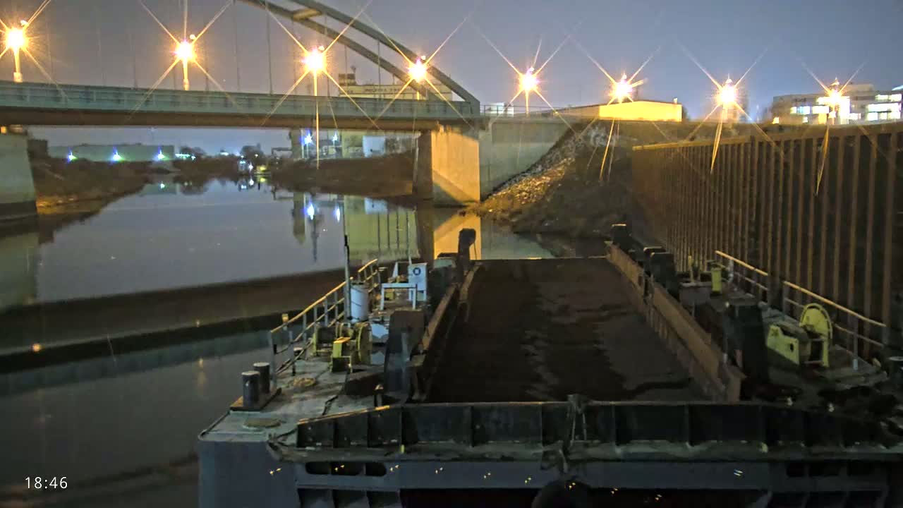 A barge laden with dark material is docked at night under a bridge with lights illuminating the waterway.