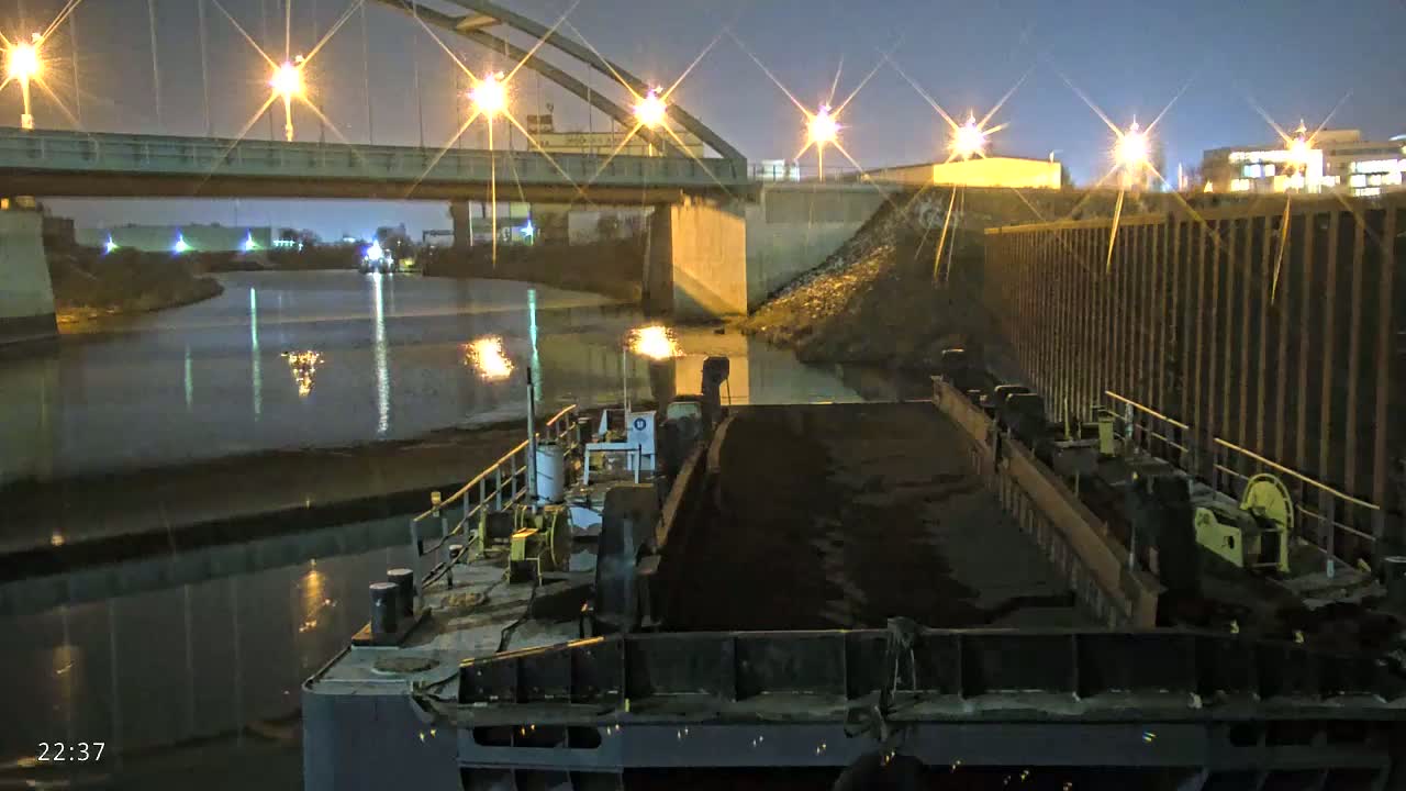 A barge filled with dark material is moored under a bridge at night, with lights illuminating the scene.