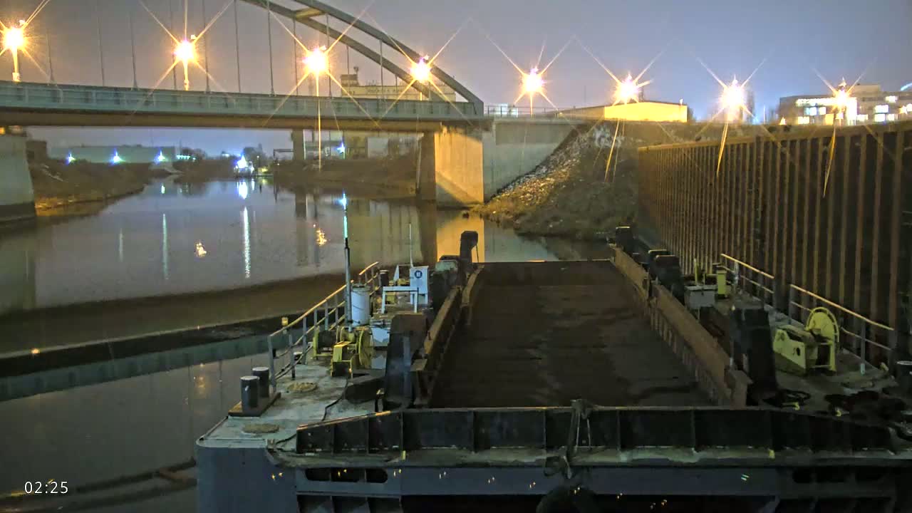 A barge carrying a dark cargo is moored at night under a bridge, illuminated by lights along the waterway.