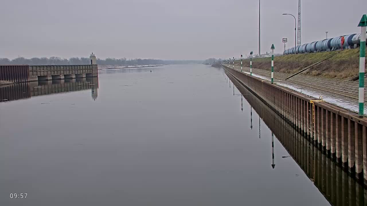 A calm, gray river is seen on an overcast day, with a concrete retaining wall and a lock on the opposite bank.