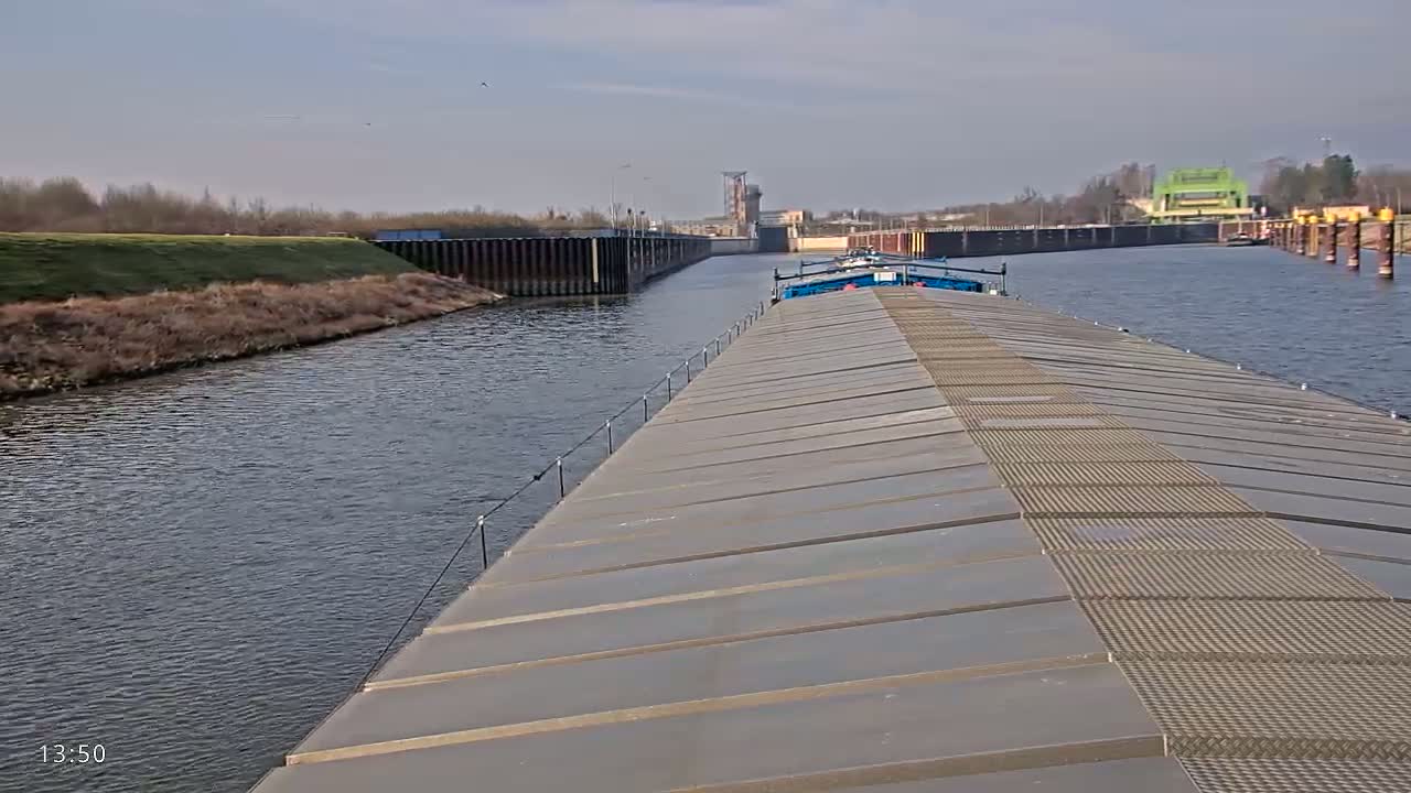 A barge travels down a canal on a partly sunny day, passing a lock and other vessels.