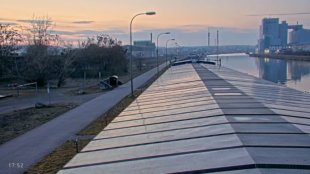 A long barge sits on a calm canal at sunrise, with a paved path and bare winter trees lining the bank.