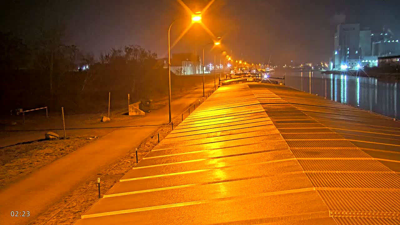 A long, illuminated barge deck at night next to a calm canal, with a path and bare trees along the bank.