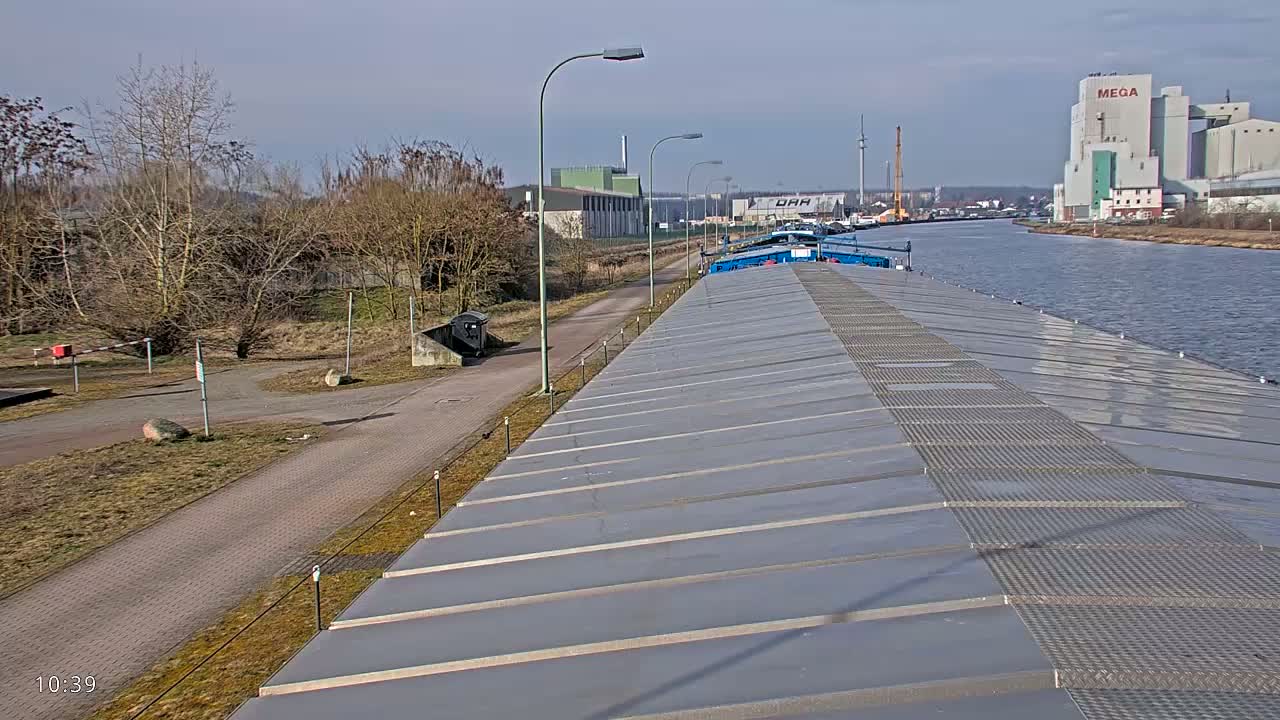 A barge's deck, under a sunny sky, overlooks a waterway with a paved path and bare trees along its bank, leading to industrial buildings in the distance.