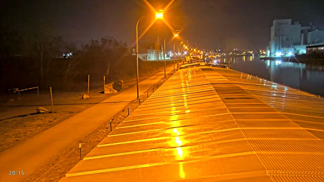 A wet, illuminated walkway stretches alongside a waterway at night, leading toward a factory and a calm river reflecting city lights.