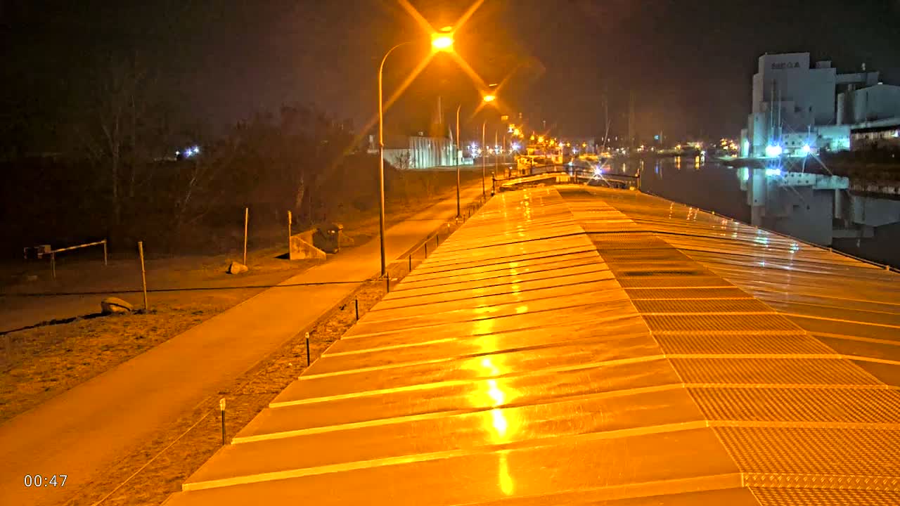 A nighttime view from atop a barge shows a paved walkway and lights along a canal, with a large industrial building reflected in the water.