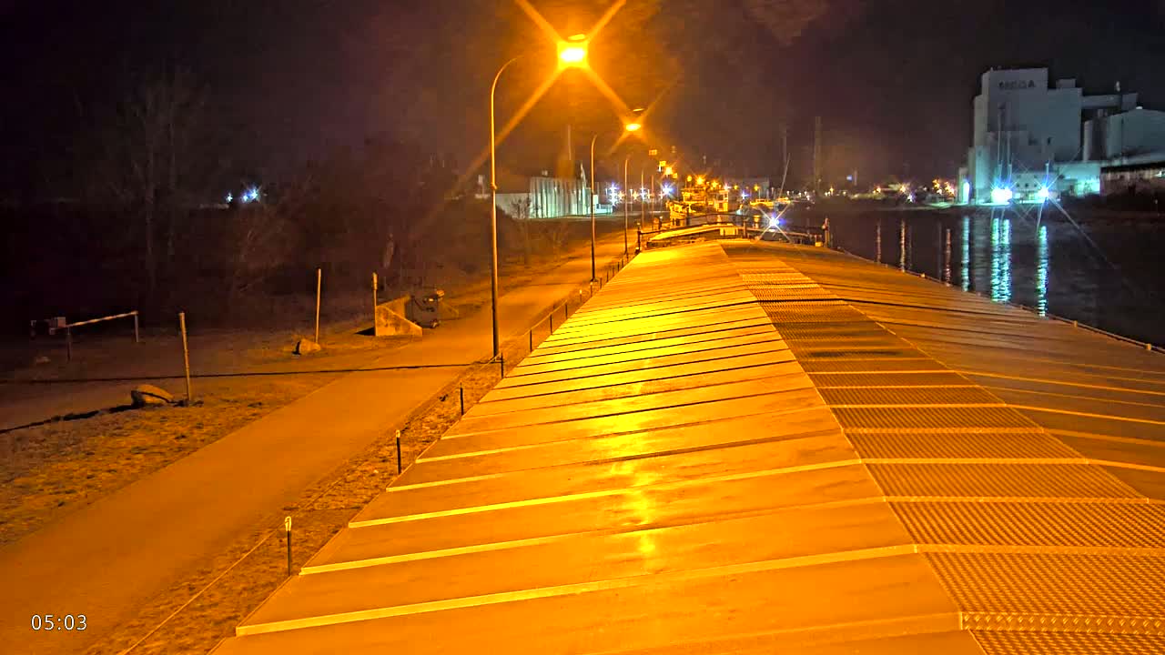 A long, illuminated barge deck stretches out at night alongside a paved path, with lights lining the path and industrial buildings visible across a calm body of water in the background.