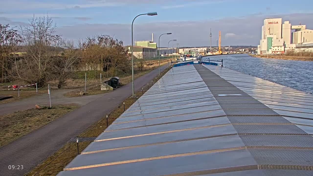 A barge travels down a canal on a partly cloudy day, passing industrial buildings and a paved path.
