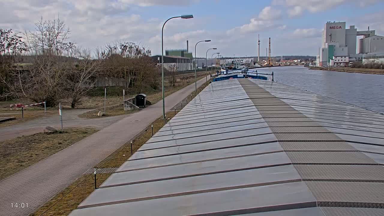 A barge's deck is seen from above, with a paved path and trees visible along a calm waterway under a partly cloudy sky.
