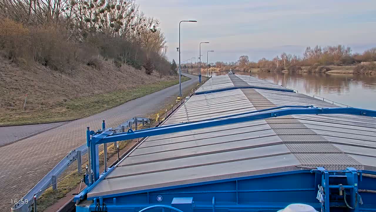 A long barge with a blue frame and gray deck travels down a calm river under a cloudy sky, passing a paved road and bare trees.