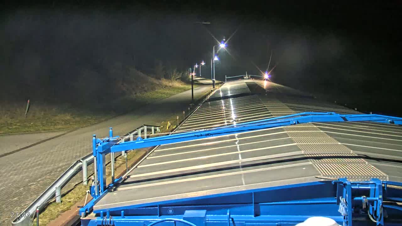 A nighttime view from atop a blue structure shows a long, flat, wet surface extending into the distance, illuminated by streetlights lining a paved path beside a sloped bank.