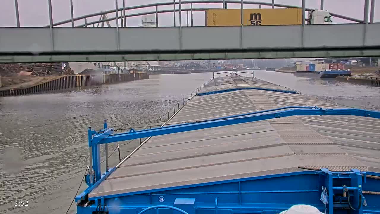 A blue barge travels on a waterway under an overcast sky, passing under a bridge.